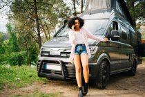 Ernste junge Afroamerikanerin spricht mit Smartphone und gestikuliert, während sie sich an ein im Wald geparktes Wohnmobil lehnt — Stockfoto