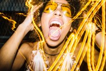 Expressive young ethnic lady with Afro hair in stylish sunglasses and top touching head and screaming loudly while chilling in nightclub near freeze lights — Stock Photo