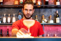 Fröhlicher Barmann serviert Glas mit Alkoholcocktail auf Theke in Bar und blickt in die Kamera — Stockfoto