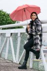 Vista lateral da fêmea étnica otimista com guarda-chuva sorrindo e olhando para longe enquanto se inclina no corrimão da ponte no dia chuvoso no parque — Fotografia de Stock