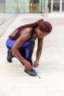 Donna atletica etnica seduta sulla strada e stringendo i lacci delle scarpe da ginnastica — Foto stock
