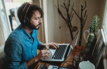 Side view of young man in headphones using synthesizer and laptop at table at home — Stock Photo