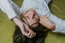From above of young green eyed female with long fair hair wearing white blouse touching head with eyes closed lying on green fabric — Stock Photo