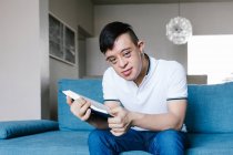 Baixo ângulo de adolescente étnico com síndrome de Down lendo livro interessante enquanto sentado olhando para a câmera no sofá na sala de estar em casa — Fotografia de Stock