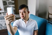 Улыбающийся латиноамериканец с синдромом Дауна, сидящий дома на диване и стреляющий в себя на смартфоне — стоковое фото