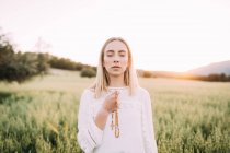 Femme fidèle en robe blanche tenant des perles avec croix tout en priant dans la solitude sur un terrain rural calme dans la nature — Photo de stock