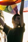 Зачарований чоловічий гей, що стоїть з закритими очима, підняв веселку ЛГБТ прапор під час заходу сонця в місті — стокове фото