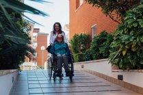 Glückliche erwachsene Tochter schiebt Rollstuhl mit älterer Mutter beim Spaziergang an sonnigem Tag — Stockfoto