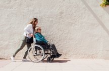 Vista lateral da filha adulta empurrando cadeira de rodas com a mãe sênior enquanto caminha ao longo da rua da cidade no dia ensolarado — Fotografia de Stock