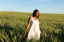 Sonriente joven dama negra en vestido de verano blanco paseando por el campo de trigo verde mientras mira a la cámara durante el día bajo el cielo azul - foto de stock