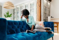 Jeune femme noire en tenue décontractée avec écouteurs assis sur un canapé bleu confortable à la maison et le magazine de lecture tout en profitant de temps libre à la maison — Photo de stock