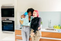 Радостные стильные многорасовые подруги в повседневной одежде стоят на домашней кухне и делают селфи с помощью мобильного телефона — стоковое фото