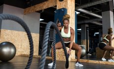 Fuerte atleta afroamericana en ropa deportiva haciendo ejercicio con cuerdas de batalla mientras mira hacia adelante durante el entrenamiento de alta intensidad en el gimnasio - foto de stock