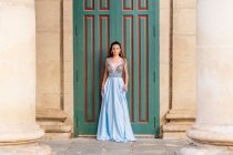 Cuerpo completo de encantadora modelo femenino joven en elegante vestido de noche azul de moda de pie cerca de la entrada del edificio envejecido y mirando a la cámara - foto de stock