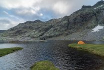 Tenda arancione situato sulla riva erbosa del lago Laguna Grande contro Sierra de Gredos catena montuosa e cielo nuvoloso in Avila, Spagna — Foto stock