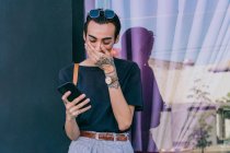 Bajo ángulo de mensajería masculina gay en las redes sociales en el teléfono móvil mientras está de pie en la calle y sonriendo - foto de stock