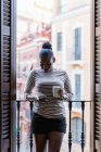 African American female with cup of hot drink surfing internet on cellphone between shutters at home — Stock Photo