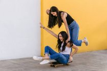 Fröhlicher Teenager mit gekreuzten Beinen fährt Skateboard mit zufriedenen weiblichen Geschwistern tagsüber auf Gehweg — Stockfoto