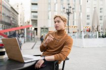 Потрясающий азиатский фрилансер, сидящий с ноутбуком за столом в уличном кафе и рефлекторно поднимающий голову, отводя взгляд в сторону — стоковое фото
