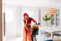 Молодая креативная дизайнерша в модном наряде и очках разговаривает по смартфону, стоя с велосипедом в современной светлой квартире — стоковое фото