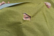Jeune femelle aux yeux verts méconnaissable regardant à travers un trou déchiré dans un tissu vert — Photo de stock