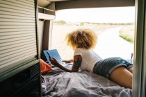 Young African American female traveler with curly hair working on laptop while lying down inside camper van during summer holidays — Stock Photo
