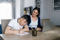 Adolescente étnico com síndrome de Down desenhando com lápis no papel enquanto se senta à mesa com freelancer feminino trabalhando no laptop em casa — Fotografia de Stock