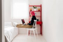 Side view of focused young female freelancer in casual clothes sitting on chair and using laptop while working on project in light modern apartment — Stock Photo