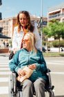 Adult woman pushing wheelchair with senior mother and crossing road in city during stroll in summer — Stock Photo