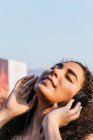 Беззаботная молодая латиноамериканка с вьющимися волосами слушает музыку через наушники, расслабляясь на улице в солнечный летний вечер. — стоковое фото