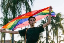Content gay male walking with rainbow LGBT flag in raised hands along street in tropical city — Stock Photo