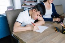 Ethnic teenage boy with Down syndrome drawing with pencils on paper while sitting at table with cropped unrecognizable female freelancer working on laptop at home — Stock Photo