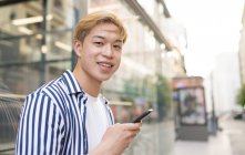 Conteúdo macho étnico com cabelo loiro em pé na rua e navegando telefone celular enquanto olha para a câmera — Fotografia de Stock