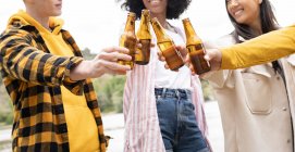 Cropped unrecognizable man and women clinking bottles of beer and proposing toast while resting on shore of river in nature — Stock Photo