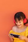 Контент ребенка серфинг интернет на мобильном телефоне во время прослушивания песни из беспроводной гарнитуры на оранжевом фоне — стоковое фото