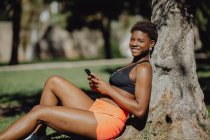 Fröhliche Afroamerikanerin in Sportkleidung hält Smartphone in der Hand, während sie an sonnigen Tagen im Gras sitzt — Stockfoto
