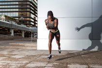 Vista laterale dell'atleta afroamericana che salta in avanti mentre corre vicino al moderno muro di cinta sulla strada della città — Foto stock
