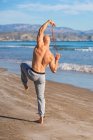 Задний вид неузнаваемого спортсмена без рубашки, протягивающего руки с эластичной лентой, стоя на одной ноге, тренирующегося на пустом солнечном пляже — стоковое фото