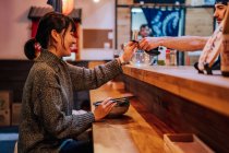 Vista lateral de la mujer asiática satisfecha en suéter sonriendo mientras toma cuchara de trabajador sentado en el mostrador de madera en el bar de ramen - foto de stock