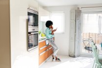Vista lateral da jovem afro-americana em uso casual comer maçã e navegar na internet no telefone celular enquanto está em casa cozinha — Fotografia de Stock
