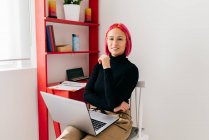 Freelancer jovem focado em roupas casuais sentado na cadeira olhando para a câmera usando laptop enquanto trabalhava no projeto em luz apartamento moderno — Fotografia de Stock