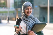 Glückliche muslimische Unternehmerin im Hijab und mit Ordner, der auf der Straße steht und auf dem Handy surft, während Geschäftsprojekt diskutiert wird — Stockfoto