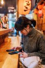 Азійська жінка у повсякденному светрі, користуючись мобільним телефоном за стійкою в традиційному барі рамена. — стокове фото