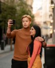Loving multiethnic couple hugging and taking self shot on smartphone while standing in street — Stock Photo