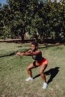 Vista lateral da jovem aptidão afro-americana fazendo agachamentos no parque em um dia ensolarado — Fotografia de Stock