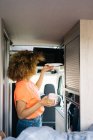 Вид збоку на невпізнавану жінку-мандрівника з кучерявим волоссям, що вкладає тарілку в мікрохвильову піч, підігріваючи їжу всередині сучасного каравана — стокове фото