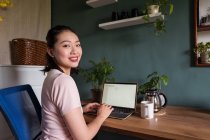 Vista lateral de la mujer asiática feliz freelancer leer documentos en el ordenador portátil mientras está sentado en la mesa durante el trabajo remoto mirando a la cámara - foto de stock