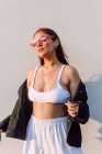 Fiduciosa giovane donna che indossa abiti casual alla moda e occhiali da sole in piedi vicino al muro bianco sotto il sole — Foto stock