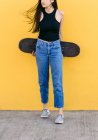 Cropped unrecognizable young female skater with skateboard standing looking away on walkway with colorful yellow wall on the background in daytime — Stock Photo