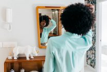 Vista posterior de una hermosa joven afroamericana en traje casual tocando el pelo rizado mientras está de pie en la habitación y mirando el espejo - foto de stock